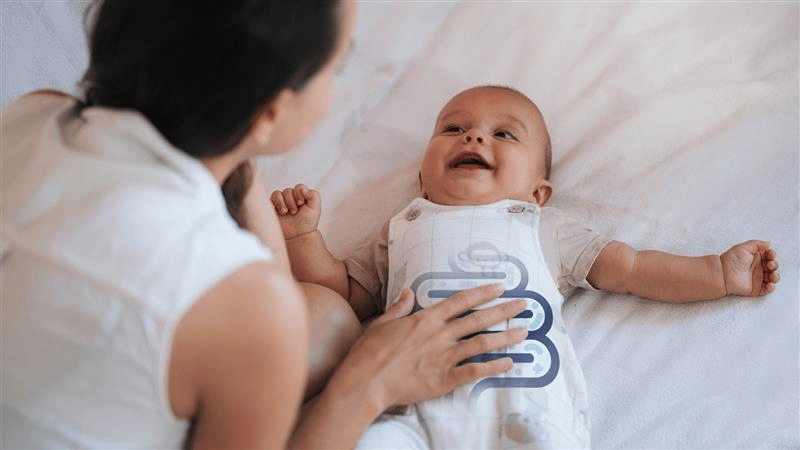 Baby lying on a bed with a person next to them, wearing a onesie with a blue design.